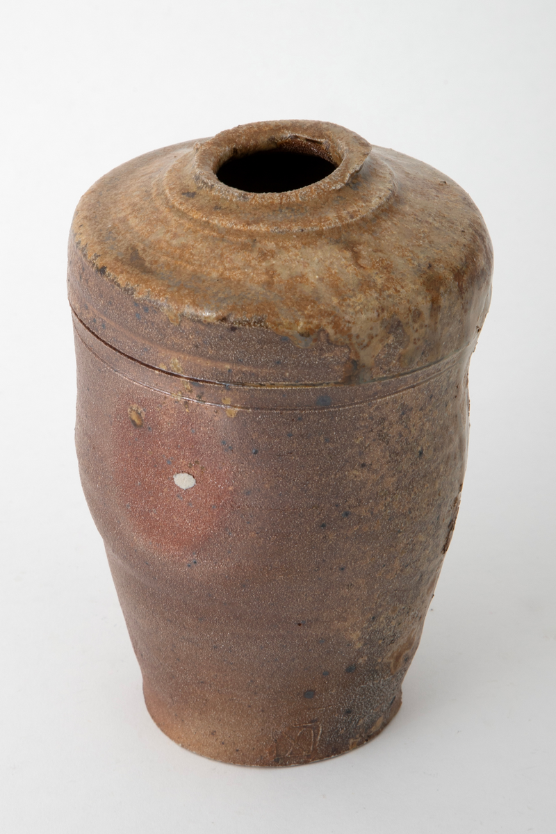 Krukke i steingods, avsmalnende nedover med uregelmessig profil. Liten åpning på svakt hvelvet, profilert topp. To ringer risset inn i godset på øvre del av korpus. Krukken er saltglasert og er brun på den ene siden og gulbrun på den andre.