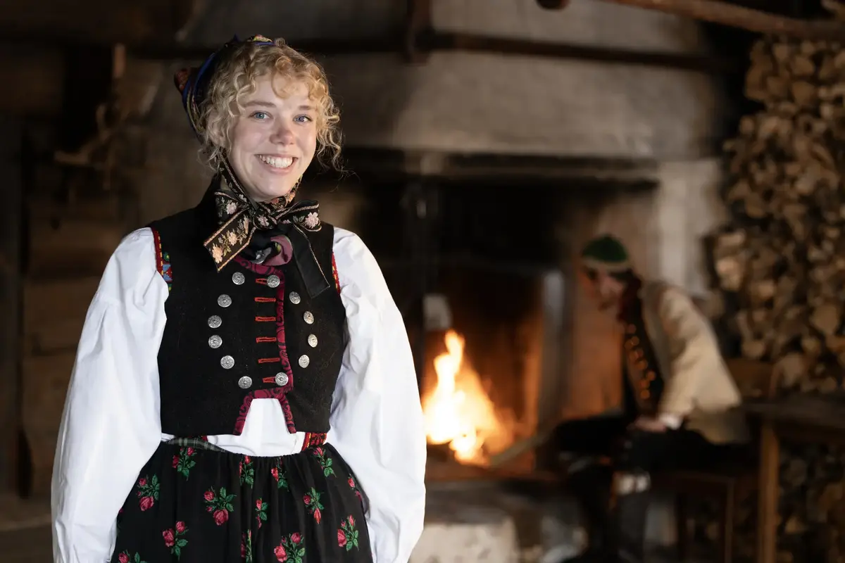 En ung kvinne i en tradisjonell norsk bunad smiler til kameraet i en gammel stue med peis og vedstabel.