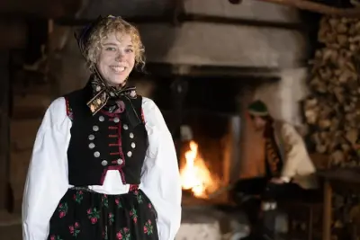 En ung kvinne i en tradisjonell norsk bunad smiler til kameraet i en gammel stue med peis og vedstabel.