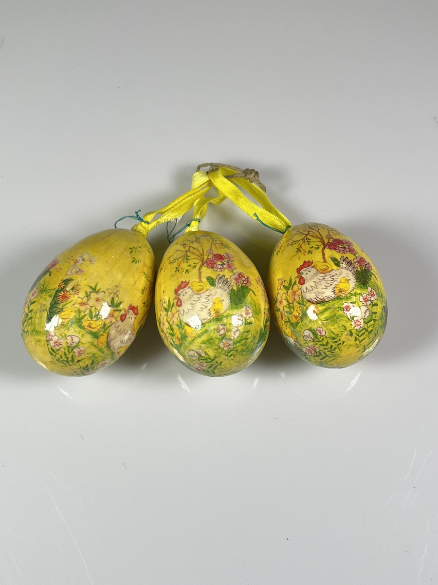 Tre st påskägg med upphängningsband. Äggen är gula med bild av en höna och kycklingar bland gräs och blommor och en vårgrönskande gren. De tre äggen är sammanbundna med ett grönt papperssnöre.

Från Arne Ekströms handelsmuseum.
