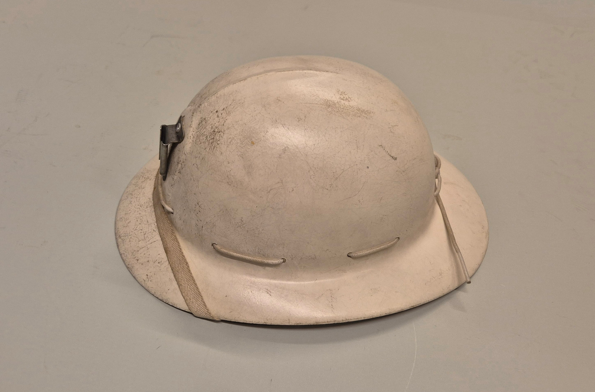 Hjelm i hvit plast med brem. Lampefeste foran på hjelmen. Innmaten utgjøres av lær og bomullsbånd, og hjelmen har et ca 1 cm bredt bomullsbånd til feste under haka. Alt er festet i en hvit rund snor som går rundt hele hjelmen.