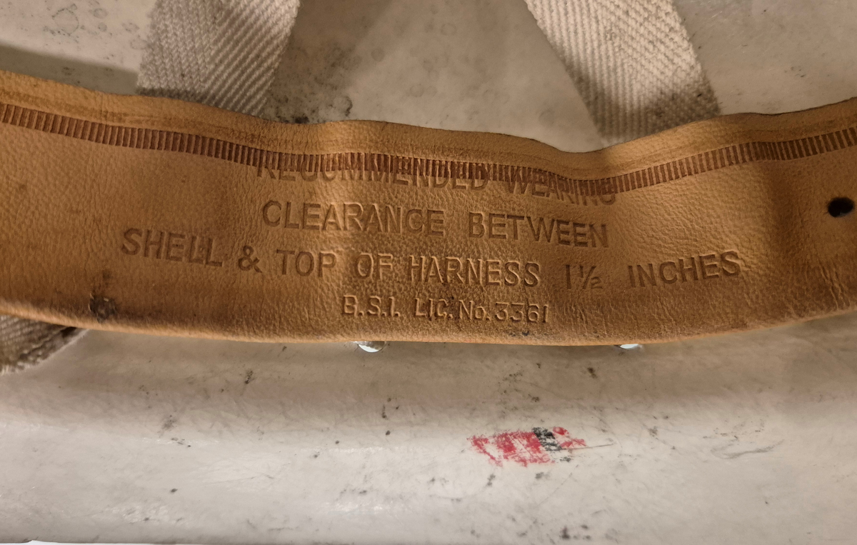 Hjelm i hvit plast med brem. Lampefeste foran på hjelmen. Innmaten utgjøres av lær og bomullsbånd, og hjelmen har et ca 1 cm bredt bomullsbånd til feste under haka. Alt er festet i en hvit rund snor som går rundt hele hjelmen.