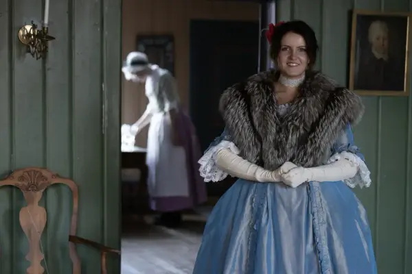 En kvinne i staselig, blå 1700-tallskjole. En annen kvinne kledd som en tjenestejente står i bakgrunnen. 