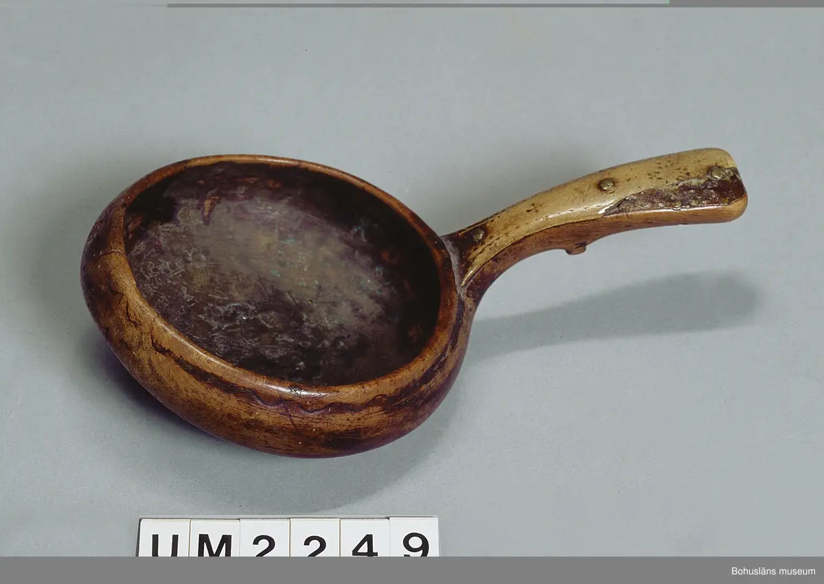 Rund skålformad kåsa med handtag skuret ur ett stycke. Mynningskanten är något indragen och mittemot handtaget är kanten upphöjd.
På utsidan kanten löper en ristad vågformad dekor.
Skaftet är böjt och bredare baktill. Ovansidan har en hornbit fastsatt med mässingsnubb. Hornbiten har ristad dekor med vågformad kant samt ett litet kors och en större korsliknande dekor.
På handtagets undersida finns ett tvärgående, utstickande parti troligtvis för att kunna hålla den lättare eller för att vila den på ett kärls kant.
Upptill på handtagets undersida finns ett inristat bomärke.
En bit av hornbiten är mörkare och sekundärt ilagad.
Kåsan har en spricka i kantens upphöjning. Inuti är det ärggröna fläckar.
Tre små hål, troligtvis efter häftstift.

Uppgifter i samband med besök från Ájtte, Svenskt Fjäll- och Samemuseum, Jokkmokk:
Lulesamisk, låg modell. Ägarnas bomärke

Ur handskrivna katalogen 1957-1958:
Träskål
Diam.: 8,5. Skopa el. Slev av trä. Föremålet helt. Lappland

Lappkatalog: 7