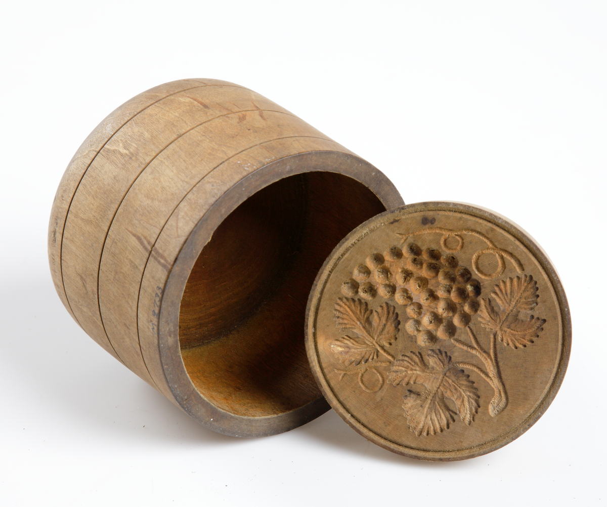 Smörpress av trä med lock. Cylindrisk. Skuren dekor i lockets undersida.
Inskrivet i huvudkatalog 1904.