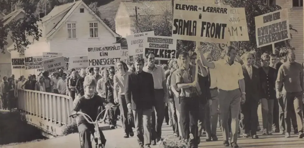 Ei gruppe demonstrantar går gjennom eit lite sentrumsområde og held oppe banner om nynorske lærebøker i skulen