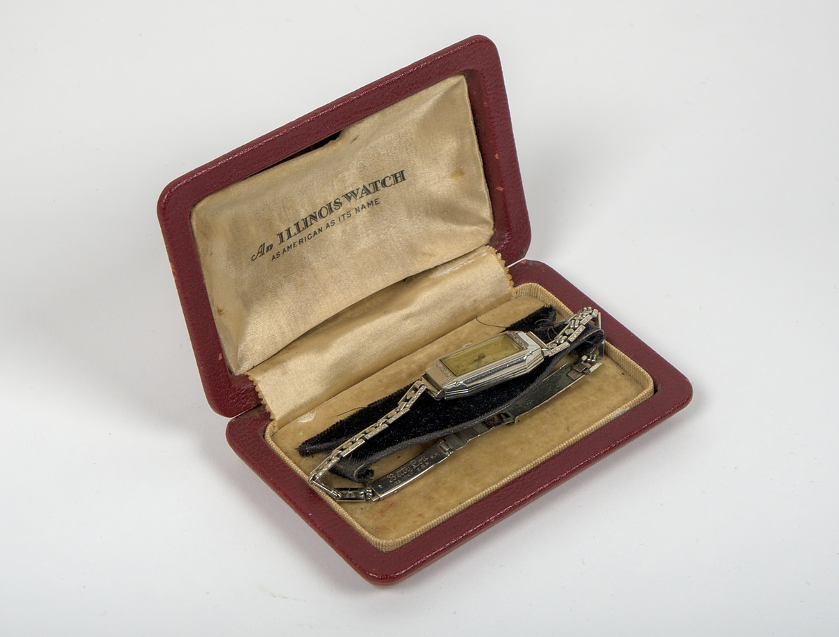 Armbandsur med tillhörande etui.

a) Damarmbandsur i art deco-stil med rektangulärt boett och länk i vitmetall. Urtavla med avlångt kvadratisk form, med arabiska siffror och två visare. Länken består av dekorerade länkar med blomformade detaljer. Boettens baksida graverad “Edith 1931”. På insidan av länken graverat “Betty Lou”, samt stämplat “1/20 12K G.F.” och “E.B.R.”.

Stämpeln “12K G.F.” antyder att ytskiktet består av 12 karats (vitt) guld, så kallad gold filled-metall.

b) Etui med kvadratisk form och högt vält lock. Utsidan klädd i röd läderimitation av formpressad plast. Locket är på insidan fodrat med beige siden och bär texten: "An Illinois Watch as american as its name". Insidans nederdel klädd i beige filtmaterial med svart uppbyggnad av filt för fästning av armbandsuret. Undersidan bär fodraltillverkarens stämpel: *Oläsligt i kursiv stil* följt av "Patented boxes".