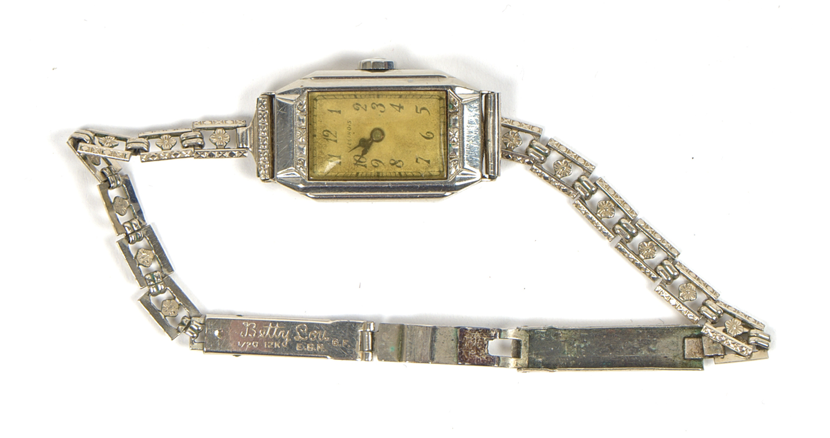 Armbandsur med tillhörande etui.

a) Damarmbandsur i art deco-stil med rektangulärt boett och länk i vitmetall. Urtavla med avlångt kvadratisk form, med arabiska siffror och två visare. Länken består av dekorerade länkar med blomformade detaljer. Boettens baksida graverad “Edith 1931”. På insidan av länken graverat “Betty Lou”, samt stämplat “1/20 12K G.F.” och “E.B.R.”.

Stämpeln “12K G.F.” antyder att ytskiktet består av 12 karats (vitt) guld, så kallad gold filled-metall.

b) Etui med kvadratisk form och högt vält lock. Utsidan klädd i röd läderimitation av formpressad plast. Locket är på insidan fodrat med beige siden och bär texten: "An Illinois Watch as american as its name". Insidans nederdel klädd i beige filtmaterial med svart uppbyggnad av filt för fästning av armbandsuret. Undersidan bär fodraltillverkarens stämpel: *Oläsligt i kursiv stil* följt av "Patented boxes".