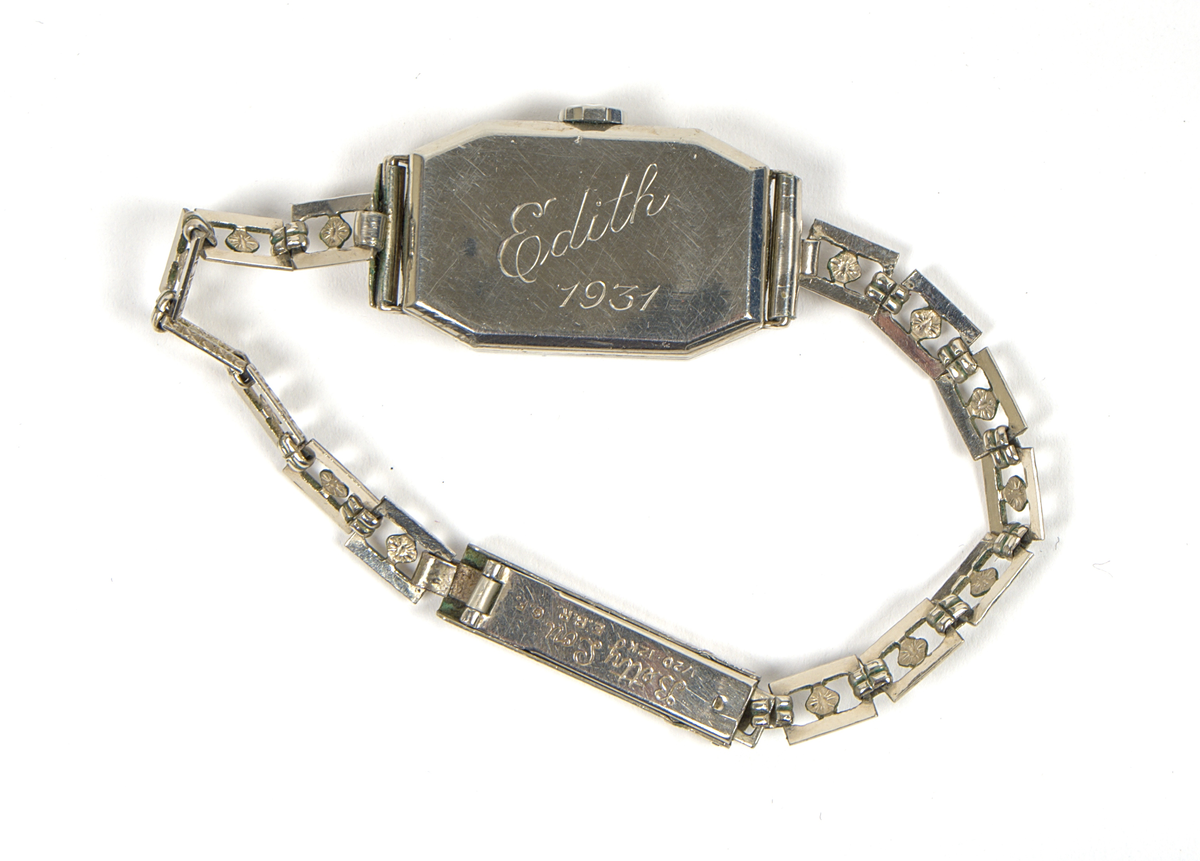 Armbandsur med tillhörande etui.

a) Damarmbandsur i art deco-stil med rektangulärt boett och länk i vitmetall. Urtavla med avlångt kvadratisk form, med arabiska siffror och två visare. Länken består av dekorerade länkar med blomformade detaljer. Boettens baksida graverad “Edith 1931”. På insidan av länken graverat “Betty Lou”, samt stämplat “1/20 12K G.F.” och “E.B.R.”.

Stämpeln “12K G.F.” antyder att ytskiktet består av 12 karats (vitt) guld, så kallad gold filled-metall.

b) Etui med kvadratisk form och högt vält lock. Utsidan klädd i röd läderimitation av formpressad plast. Locket är på insidan fodrat med beige siden och bär texten: "An Illinois Watch as american as its name". Insidans nederdel klädd i beige filtmaterial med svart uppbyggnad av filt för fästning av armbandsuret. Undersidan bär fodraltillverkarens stämpel: *Oläsligt i kursiv stil* följt av "Patented boxes".