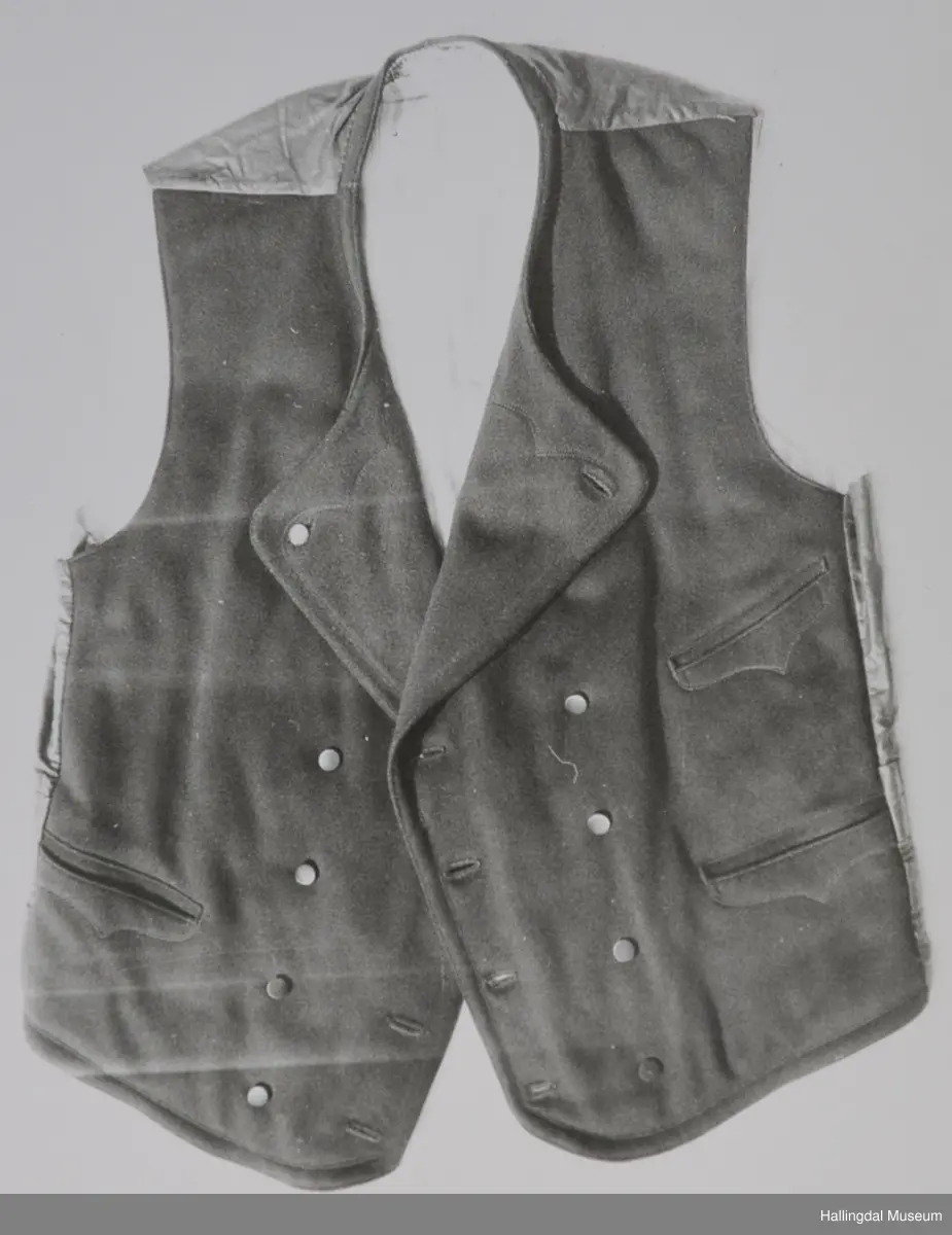 Rygg av noko slags svart, tunt, blankt bommullsty, i livet på ryggen belte av same slagt tyet. Framstykke av svart klede. Ryggen er fora av med smalstriput bomullsty, kvit botnfarge og striper i blått, raudt og brunt. Framstykke er fora med blått og kvitt rutut, tjukt bomullstoff.