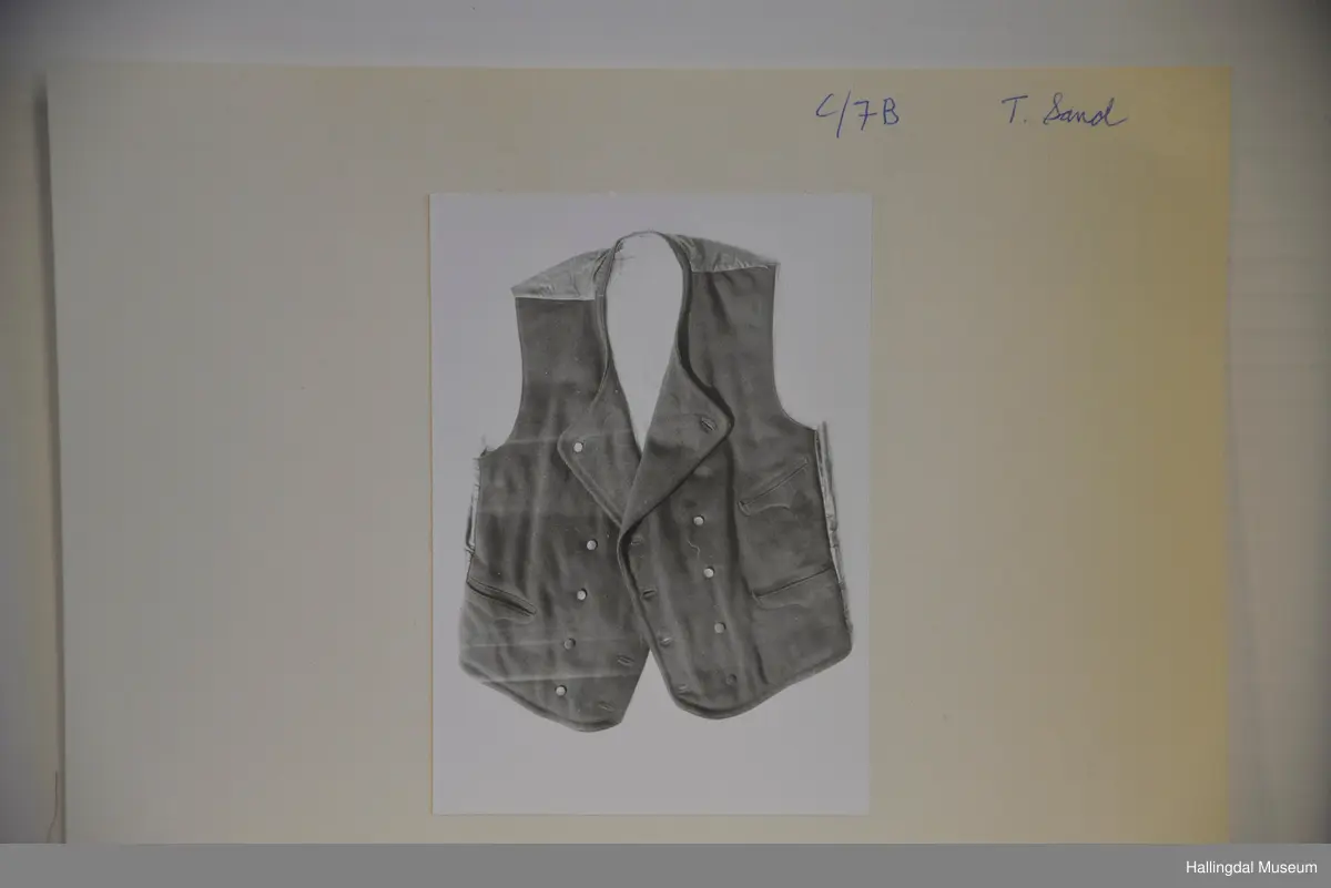 Rygg av noko slags svart, tunt, blankt bommullsty, i livet på ryggen belte av same slagt tyet. Framstykke av svart klede. Ryggen er fora av med smalstriput bomullsty, kvit botnfarge og striper i blått, raudt og brunt. Framstykke er fora med blått og kvitt rutut, tjukt bomullstoff.