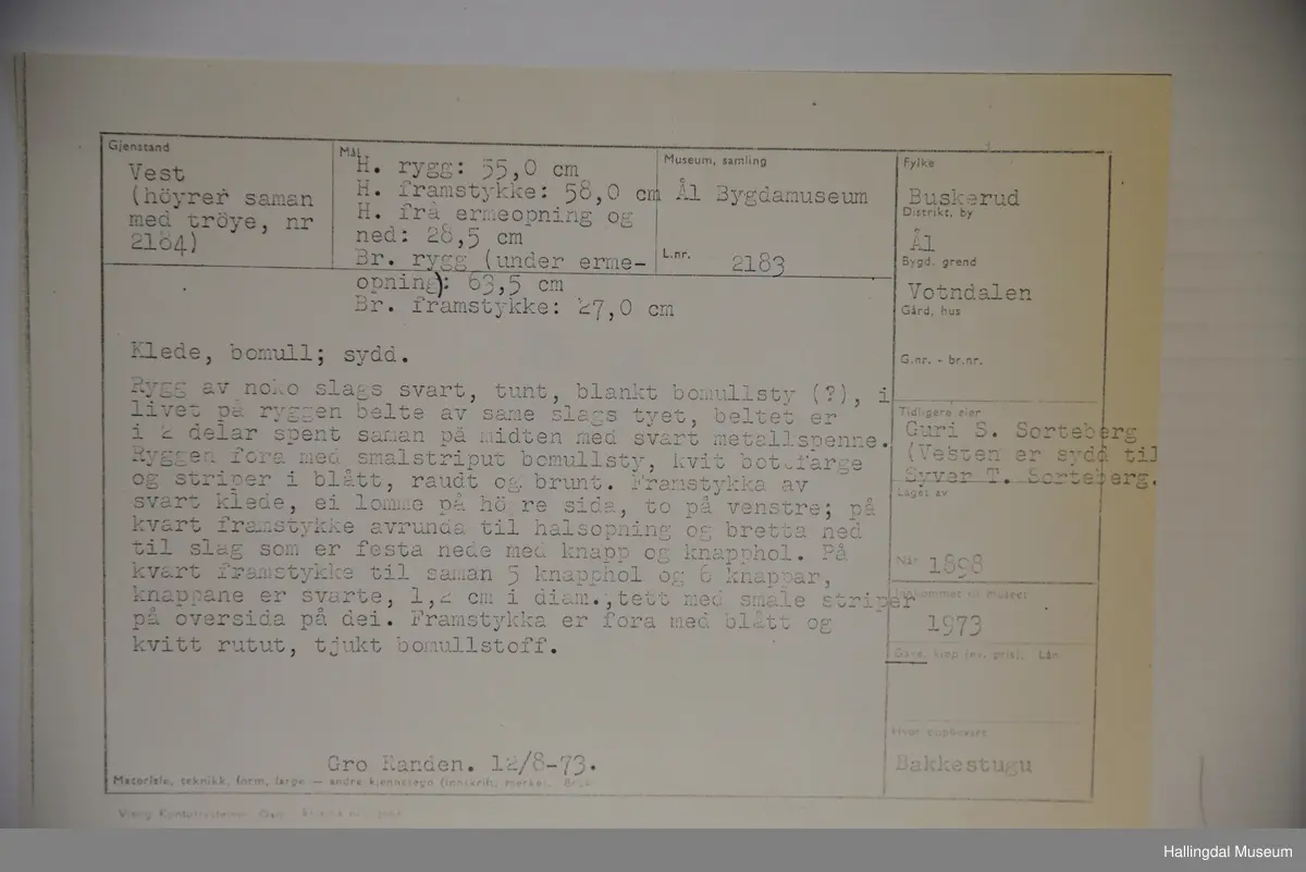 Rygg av noko slags svart, tunt, blankt bommullsty, i livet på ryggen belte av same slagt tyet. Framstykke av svart klede. Ryggen er fora av med smalstriput bomullsty, kvit botnfarge og striper i blått, raudt og brunt. Framstykke er fora med blått og kvitt rutut, tjukt bomullstoff.