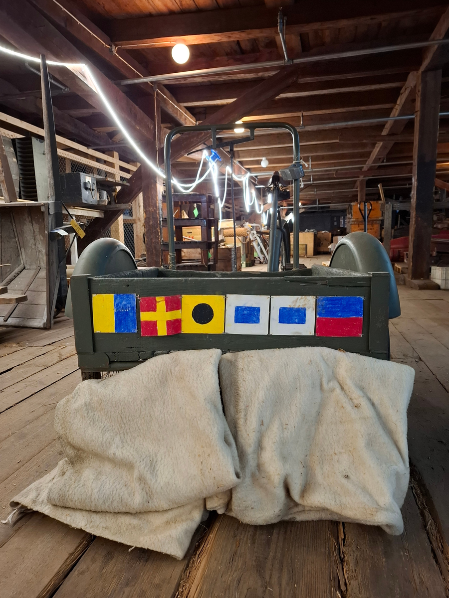 Militärgrön lådcykel av märke Monark med svarta hjul och brun lädersadel. Lådan är placerad i cykelns främre del och stabiliseras med två hjul. Styret består av en rektangulär ram monterad på lådans bakre del. Den bakre delen av cykeln ser ut som en helt vanlig cykel. 

I lådan ligger två tjocka vit-beige fleecefiltar. De båda filtarna är sydda som rektangulära påsar. I lådan finns också ett lås med texten "OPTIMUS ILCO", nyckel med texten "OPTIMUS" och kedja. 

På lådans framsida finns sex stycken handritade maritima singalflaggor på papper i olika utföranden som häftats på lådans främre del. Flaggorna ser ut att vara ritade med färgpennor/kritor. Flaggorna från vänster till höger-  
1. Gul och blå (Lodrätt):  Signalflagga K (Kilo) betyder: "Jag vill upprätta förbindelse med er".
2. Gult kryss mot röd bakgrund: Signalflagga R (Romeo) betyder: "Mitt fartyg gör ingen fart genom vattnet".
3. Gul med svart cirkel: Signalflagga I (India) betyder: "Jag ändrar min kurs babord hän".
4. Blå fyrkant mot vit bakgrund: Signalflagga S (Sierra) betyder: "Min maskin går back".
5. Blå fyrkant mot vit bakgrund: Signalflagga S (Sierra) betyder: "Min maskin går back"
6. Blå och röd (vågrätt): Signalflagga E (Echo) betyder: "Jag ändrar kurs styrbord hän".
Bokstäverna stavar tillsammans ordet "KRISSE".

Alla hjul lådcykeln behöver pumpas/lagas. Lådan i trä är också i någerlunda kritiskt skick.