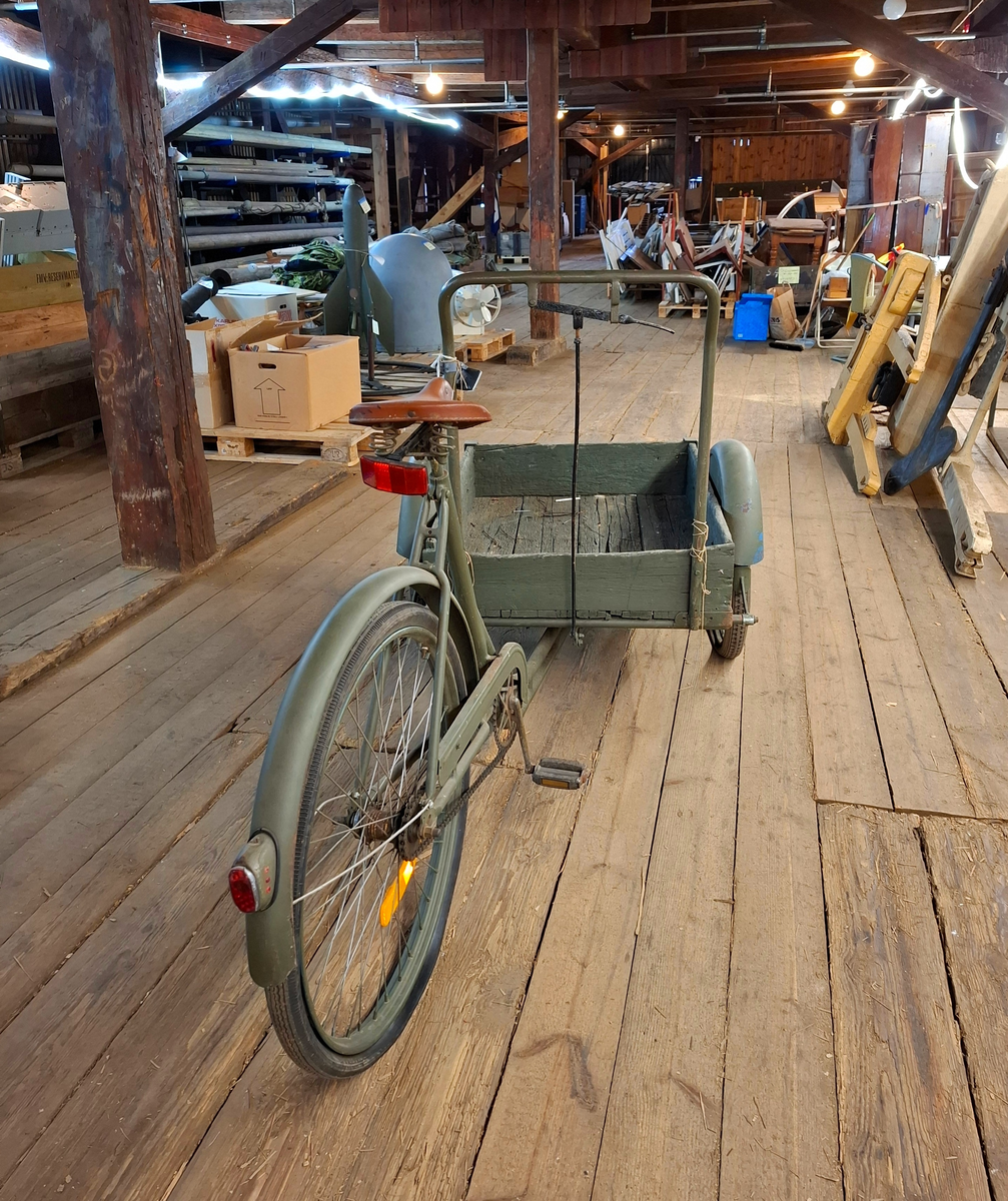 Militärgrön lådcykel av märke Monark med svarta hjul och brun lädersadel. Lådan är placerad i cykelns främre del och stabiliseras med två hjul. Styret består av en rektangulär ram monterad på lådans bakre del. Den bakre delen av cykeln ser ut som en helt vanlig cykel. 

I lådan ligger två tjocka vit-beige fleecefiltar. De båda filtarna är sydda som rektangulära påsar. I lådan finns också ett lås med texten "OPTIMUS ILCO", nyckel med texten "OPTIMUS" och kedja. 

På lådans framsida finns sex stycken handritade maritima singalflaggor på papper i olika utföranden som häftats på lådans främre del. Flaggorna ser ut att vara ritade med färgpennor/kritor. Flaggorna från vänster till höger-  
1. Gul och blå (Lodrätt):  Signalflagga K (Kilo) betyder: "Jag vill upprätta förbindelse med er".
2. Gult kryss mot röd bakgrund: Signalflagga R (Romeo) betyder: "Mitt fartyg gör ingen fart genom vattnet".
3. Gul med svart cirkel: Signalflagga I (India) betyder: "Jag ändrar min kurs babord hän".
4. Blå fyrkant mot vit bakgrund: Signalflagga S (Sierra) betyder: "Min maskin går back".
5. Blå fyrkant mot vit bakgrund: Signalflagga S (Sierra) betyder: "Min maskin går back"
6. Blå och röd (vågrätt): Signalflagga E (Echo) betyder: "Jag ändrar kurs styrbord hän".
Bokstäverna stavar tillsammans ordet "KRISSE".

Alla hjul lådcykeln behöver pumpas/lagas. Lådan i trä är också i någerlunda kritiskt skick.