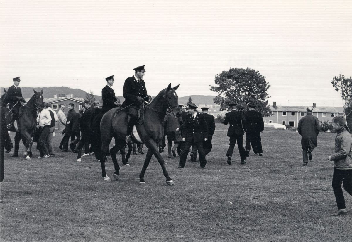 Politifolk og politiryttere på en åpen plass. Publikum. Røyksignal? Trolig en oppvisning e.l.