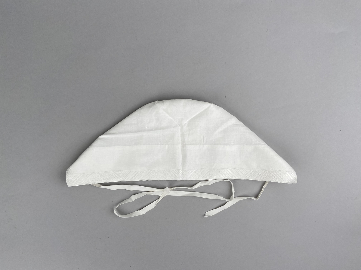 Hätta sk. fruhatt till Leksandsdräkten. Av vitt linne. Broderi i vitt på vitt och nuggor längs framkanten. Broderi vitt på vitt mitt back i nacken över sömmen.  Snörning genom hål i nederkanten. Knyts ihop med bomullsband i nacken.