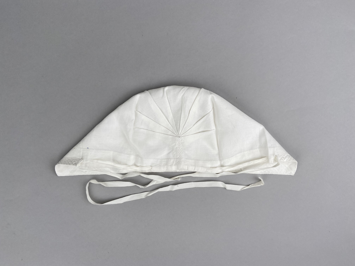 Hätta sk. fruhatt till Leksandsdräkten. Av vitt linne. Broderi i vitt på vitt och nuggor längs framkanten. Broderi vitt på vitt mitt back i nacken över sömmen.  Snörning genom hål i nederkanten. Knyts ihop med bomullsband i nacken.