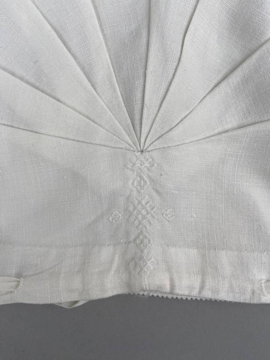 Hätta sk. fruhatt till Leksandsdräkten. Av vitt linne. Broderi i vitt på vitt och nuggor längs framkanten. Broderi vitt på vitt mitt back i nacken över sömmen.  Snörning genom hål i nederkanten. Knyts ihop med bomullsband i nacken.