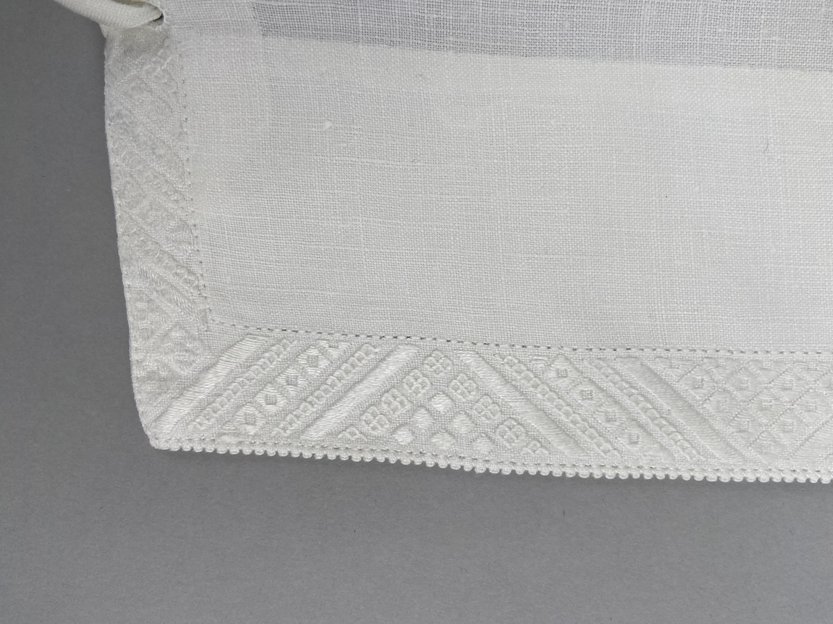 Hätta sk. fruhatt till Leksandsdräkten. Av vitt linne. Broderi i vitt på vitt och nuggor längs framkanten. Broderi vitt på vitt mitt back i nacken över sömmen.  Snörning genom hål i nederkanten. Knyts ihop med bomullsband i nacken.