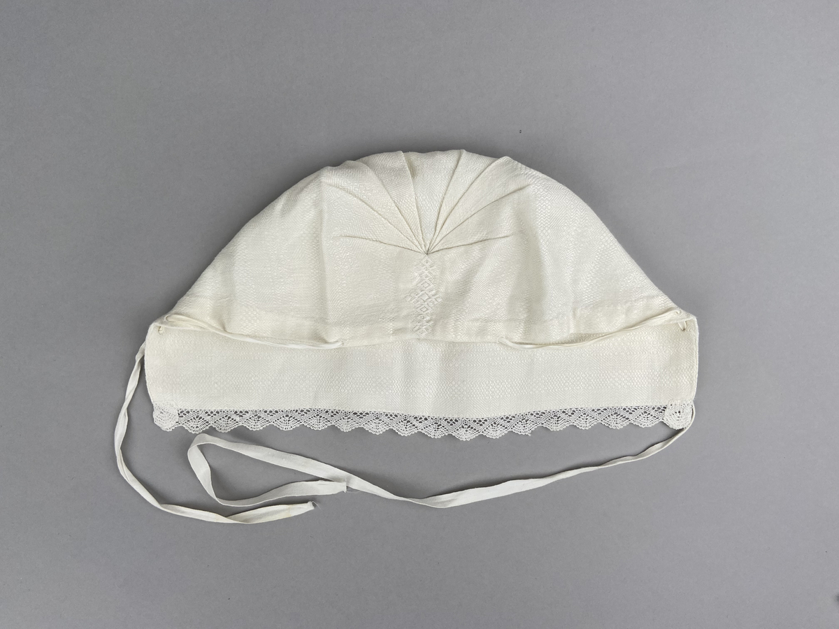 Hätta sk. fruhatt till Leksandsdräkten. Av vitt läskrus. Kantad längs framkanten med handknypplad spets. Snörning genom hål i nederkanten. Knyts ihop med bomullsband i nacken. Vitbroderi över sömmen bak.