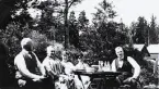 En grupp människor som sitter utomhus i en trädgård och fikar, man skönjer en skog och några byggnader i bakgrunden. Text i album: 
