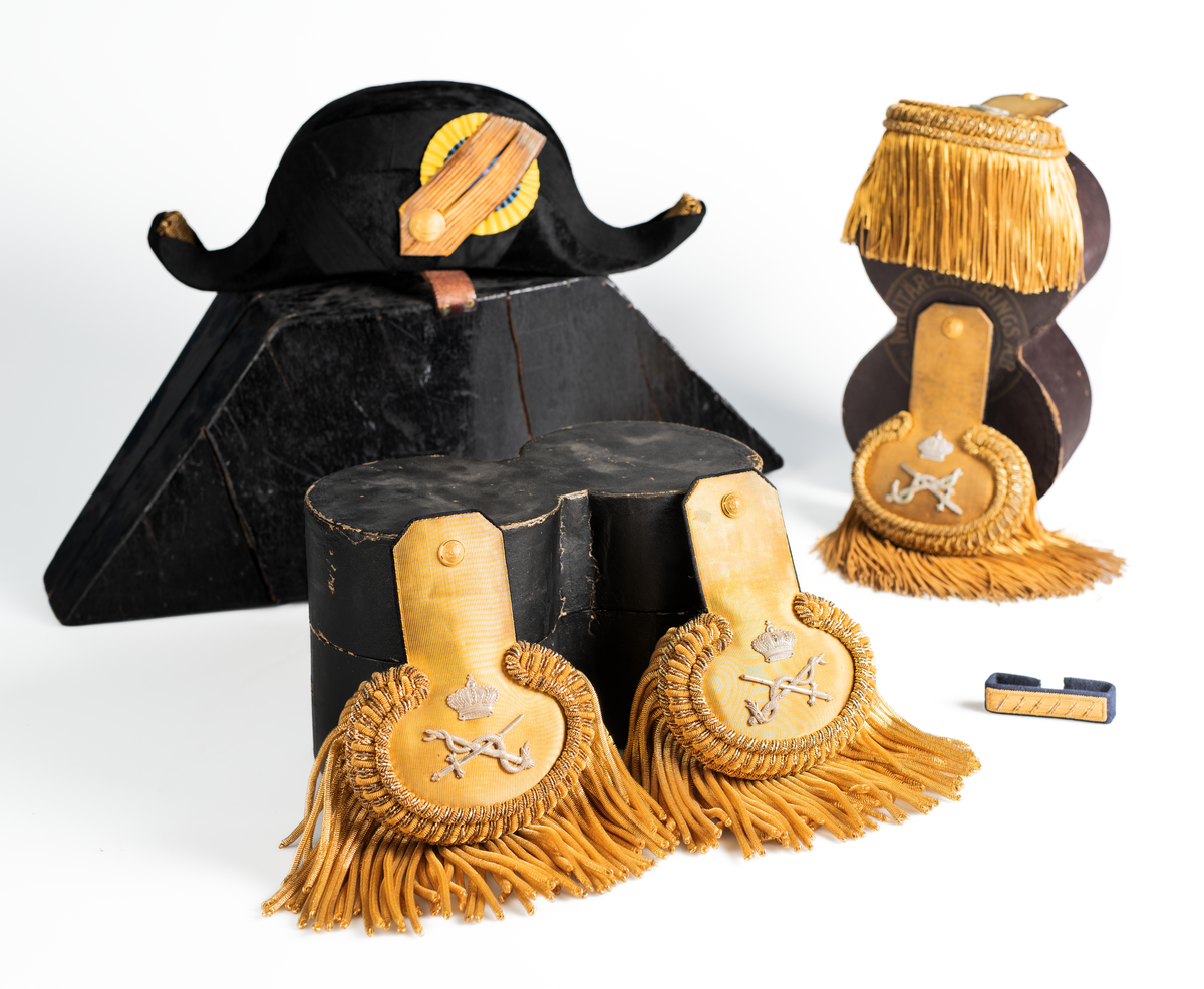 Ett par epåletter i pappask som hör till uniformen som användes av marinläkaren Anders Rudolf Prag.
Anders Prag kom tili Karlskrona 1929 som marinläkare, dispensärläkare och privatpraktiserande läkare, där hans största intresse alltid varit pediatriken, hans huvudintresse och specialitet. Anders Prag har här i staden gjort ett verkligt pionjärarbete i det att han startat barnavårdscentralsverksamheten, om vilken han aven hallit föreläsningar i Karlskrona läkareforening. I föreningen har han varit sekreterare och ordförande. Som marinläkare har Anders nått l:e marinläkargraden. De gåger han underhillit kollegerna med sång till luta, då hans sonora stamma klingat så vackert har varit verkliga högtidsstunder (ur Karlskrona Lakareförening 120 år Från hot om prygelstraff till fastställd arbetstid, SVEN SVENNERUD och HOLGER HOLMGREN 1978). "Gradbeteckningar med olikfärgat kläde mellan galonerna för civilmilitära tjänstemän med specialutbildning inom marinen; rött för marinläkare, vitt för marinintendent och lila för mariningenjör." S. 23 Flottans sjöofficersuniformer i Sjöhistoriska museets samlingar. Nils-Erik Melinder. Sida 38, samma skrift: "Från år 1937, utformades den för sjöofficeraren runda öglan till en rundad triangulär ögla, dessutom poängterad med olikfärgat kläde mellan galonerna. Statusen blev civilmilitär. Ett sammanhängande "öknamn" för den officerskategorin blev därför "päronklubben."