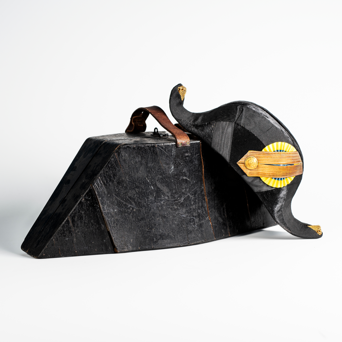 Båthatt i hattask av trä som hör till uniformen som användes av marinläkaren Anders Rudolf Prag.
Anders Prag kom tili Karlskrona 1929 som marinläkare, dispensärläkare och privatpraktiserande läkare, där hans största intresse alltid varit pediatriken, hans huvudintresse och specialitet. Anders Prag har här i staden gjort ett verkligt pionjärarbete i det att han startat barnavårdscentralsverksamheten, om vilken han aven hallit föreläsningar i Karlskrona läkareforening. I föreningen har han varit sekreterare och ordförande. Som marinläkare har Anders nått l:e marinläkargraden. De gåger han underhillit kollegerna med sång till luta, då hans sonora stamma klingat så vackert har varit verkliga högtidsstunder (ur Karlskrona Lakareförening 120 år Från hot om prygelstraff till fastställd arbetstid, SVEN SVENNERUD och HOLGER HOLMGREN 1978). "Gradbeteckningar med olikfärgat kläde mellan galonerna för civilmilitära tjänstemän med specialutbildning inom marinen; rött för marinläkare, vitt för marinintendent och lila för mariningenjör." S. 23 Flottans sjöofficersuniformer i Sjöhistoriska museets samlingar. Nils-Erik Melinder. Sida 38, samma skrift: "Från år 1937, utformades den för sjöofficeraren runda öglan till en rundad triangulär ögla, dessutom poängterad med olikfärgat kläde mellan galonerna. Statusen blev civilmilitär. Ett sammanhängande "öknamn" för den officerskategorin blev därför "päronklubben."