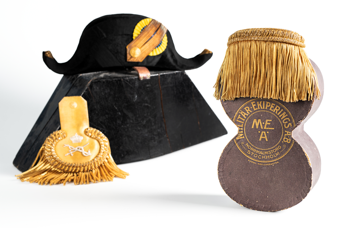 Ett par epåletter i pappask som hör till uniformen som användes av marinläkaren Anders Rudolf Prag.
Anders Prag kom tili Karlskrona 1929 som marinläkare, dispensärläkare och privatpraktiserande läkare, där hans största intresse alltid varit pediatriken, hans huvudintresse och specialitet. Anders Prag har här i staden gjort ett verkligt pionjärarbete i det att han startat barnavårdscentralsverksamheten, om vilken han aven hallit föreläsningar i Karlskrona läkareforening. I föreningen har han varit sekreterare och ordförande. Som marinläkare har Anders nått l:e marinläkargraden. De gåger han underhillit kollegerna med sång till luta, då hans sonora stamma klingat så vackert har varit verkliga högtidsstunder (ur Karlskrona Lakareförening 120 år Från hot om prygelstraff till fastställd arbetstid, SVEN SVENNERUD och HOLGER HOLMGREN 1978). "Gradbeteckningar med olikfärgat kläde mellan galonerna för civilmilitära tjänstemän med specialutbildning inom marinen; rött för marinläkare, vitt för marinintendent och lila för mariningenjör." S. 23 Flottans sjöofficersuniformer i Sjöhistoriska museets samlingar. Nils-Erik Melinder. Sida 38, samma skrift: "Från år 1937, utformades den för sjöofficeraren runda öglan till en rundad triangulär ögla, dessutom poängterad med olikfärgat kläde mellan galonerna. Statusen blev civilmilitär. Ett sammanhängande "öknamn" för den officerskategorin blev därför "päronklubben."