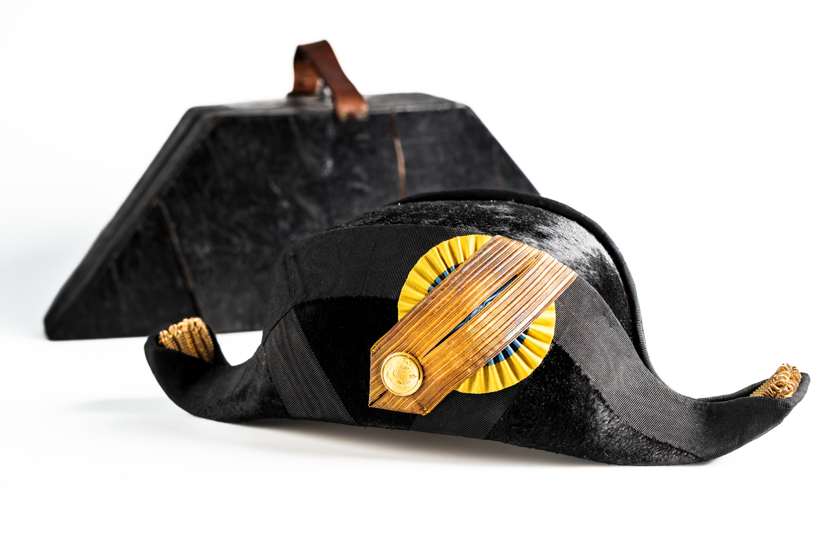 Båthatt i hattask av trä som hör till uniformen som användes av marinläkaren Anders Rudolf Prag.
Anders Prag kom tili Karlskrona 1929 som marinläkare, dispensärläkare och privatpraktiserande läkare, där hans största intresse alltid varit pediatriken, hans huvudintresse och specialitet. Anders Prag har här i staden gjort ett verkligt pionjärarbete i det att han startat barnavårdscentralsverksamheten, om vilken han aven hallit föreläsningar i Karlskrona läkareforening. I föreningen har han varit sekreterare och ordförande. Som marinläkare har Anders nått l:e marinläkargraden. De gåger han underhillit kollegerna med sång till luta, då hans sonora stamma klingat så vackert har varit verkliga högtidsstunder (ur Karlskrona Lakareförening 120 år Från hot om prygelstraff till fastställd arbetstid, SVEN SVENNERUD och HOLGER HOLMGREN 1978). "Gradbeteckningar med olikfärgat kläde mellan galonerna för civilmilitära tjänstemän med specialutbildning inom marinen; rött för marinläkare, vitt för marinintendent och lila för mariningenjör." S. 23 Flottans sjöofficersuniformer i Sjöhistoriska museets samlingar. Nils-Erik Melinder. Sida 38, samma skrift: "Från år 1937, utformades den för sjöofficeraren runda öglan till en rundad triangulär ögla, dessutom poängterad med olikfärgat kläde mellan galonerna. Statusen blev civilmilitär. Ett sammanhängande "öknamn" för den officerskategorin blev därför "päronklubben."