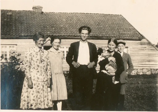 Gruppebilde framføre eit gardshus med skut, med tre kvinner, ei mann og ein gut. Kvinna lengst til høgre held ein kvalp.