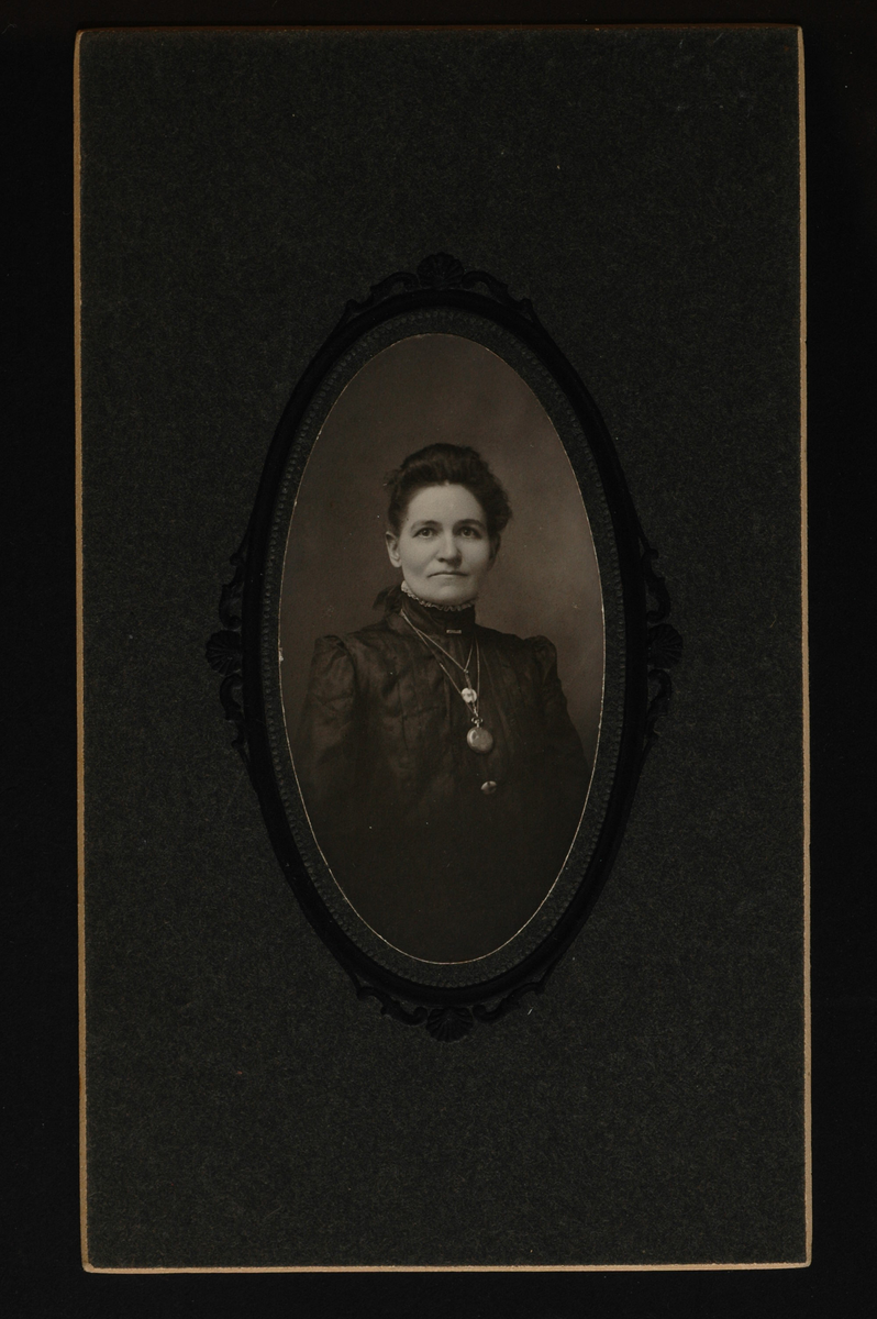 Portrett av en kvinne med mørkt hår og mørk, høyhalset overdel. Rundt halsen har hun en klokke og flere smykker.
Påskrift  bakpå: Emma Kilgore Williams