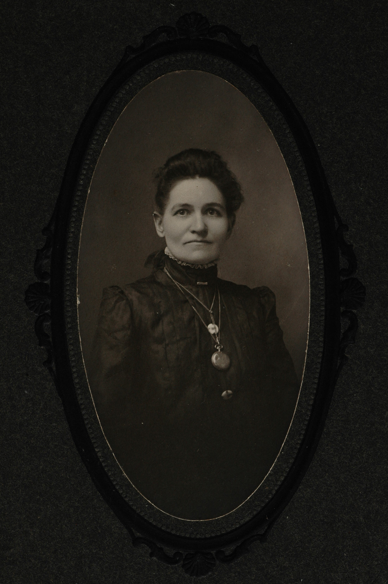 Portrett av en kvinne med mørkt hår og mørk, høyhalset overdel. Rundt halsen har hun en klokke og flere smykker.
Påskrift  bakpå: Emma Kilgore Williams