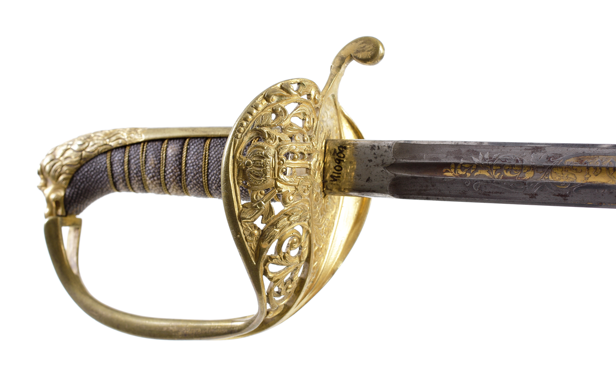 Officerssabel, M1899, med förgyllt mässingsfäste. Kaveln är klädd med svärtat fiskskinn, försedd med 8 räfflor lindade med snodd mässingstråd. Ryggbeslaget övergår i en kappa utformad som ett lejonhuvud. Parerplåten är tillverkad av gjuten mässing, enkelsidig med genombrutet kron- och bladmönster. Klingan är tveeggad, har blodrännor samt dekorationer av blomsterslingor, vapenemblem mm. På ena sidan står "Fäderneslandet" och på andra sidan "Konungen".
Baljan är tillverkad av svärtad plåt och har bygel. 

Inskriven i huvudbok år 1941.