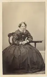 En medelålders kvinna i mörk krinolin och s.k. frumössa, sit