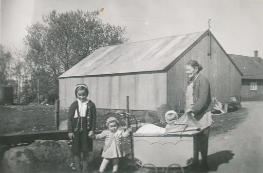 Ei kvinne med to jenter og eit mindre barn i ei barnevogn. Gardsbygningar i bakgrunnen.