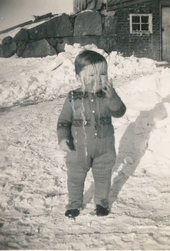 Eit lite barn ute i eit gardstun dekt av snø. I bakgrunnen ser vi ei låvebru bygd av store steinblokker. Bildet har store riper.