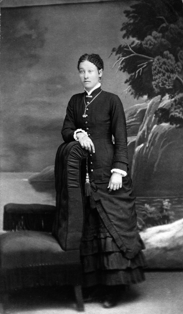 Helfigur studioportrett av en ung kvinne i mørk kjole som lener seg mot en stol.