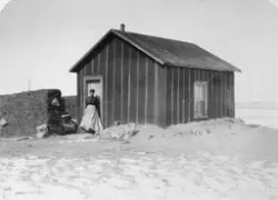 En kvinne står utenfor en enkel hytte. To vegger av torv ses