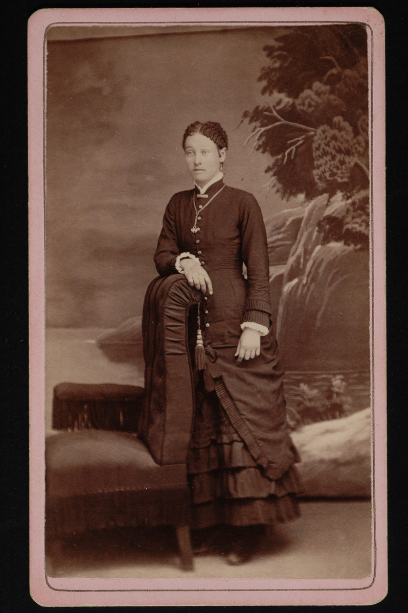 Helfigur studioportrett av en ung kvinne i mørk kjole som lener seg mot en stol.