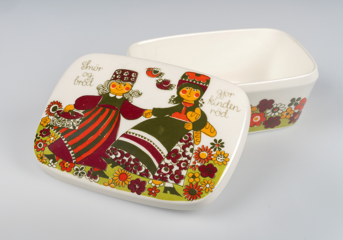 Del av servise, flintgods. Smørboks m. lokk, rektangulær form m. svakt utskrådde sider; svakt hvelvet, overfalset lokk. Utvendig dekorert m. trykkdekor (skyvebilde) med motiv av stiliserte blomster i sort konturstrek fylt med sterke farver, på lokket motiv av to kvinner i en blomstereng, den ene holder en (smør)tine og den andre har to brød under armen, rundt dem verselinjer (se innskrift). Merket på undersiden (se innskrift).