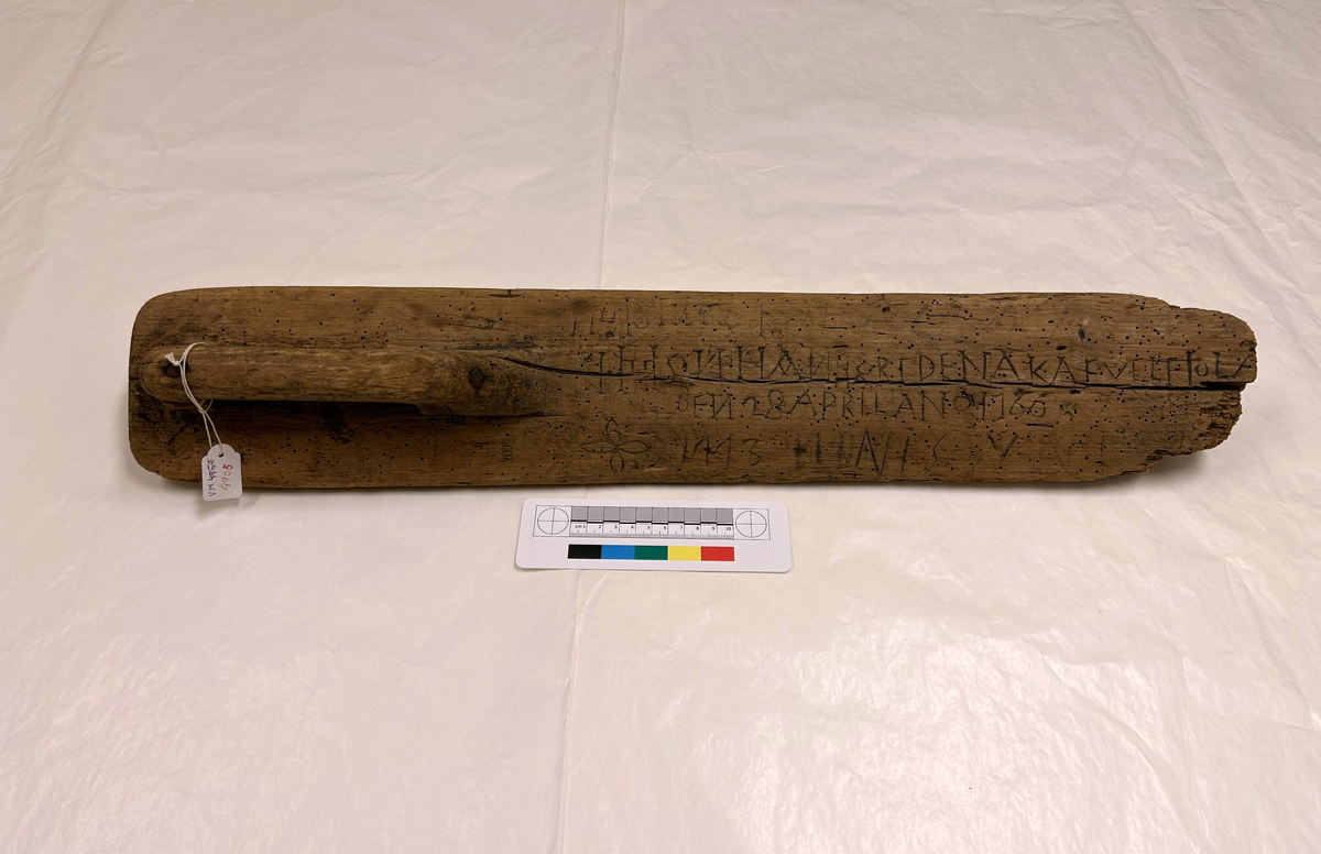 Ett rektangulärt trästycke med ett rakt utskuret handtag ur brädet som är förstärkt med spikar.
Svårtolkad inskuren text "....DENA KAFVEL -  DEN 28 APRIL ANO 1766/1773 NIS".