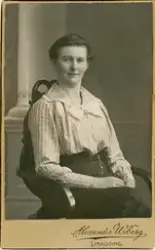 En kvinna i smalrandig blus och mörk kjol sitter i en karmst