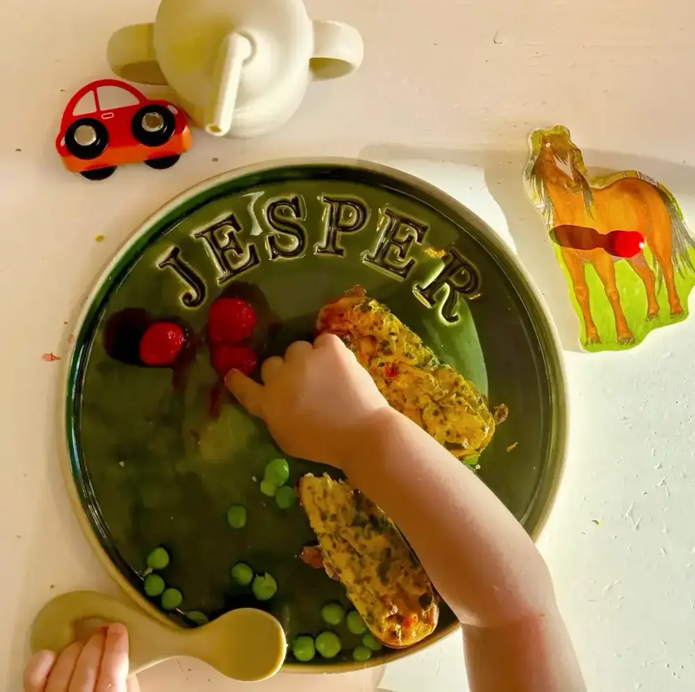 Keramisk tallrik med texten Jesper, liten barnhand äter ärtor och hallon