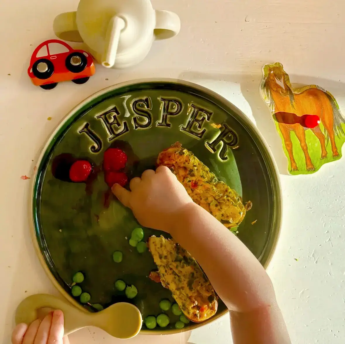 Keramisk tallrik med texten Jesper, liten barnhand äter ärtor och hallon