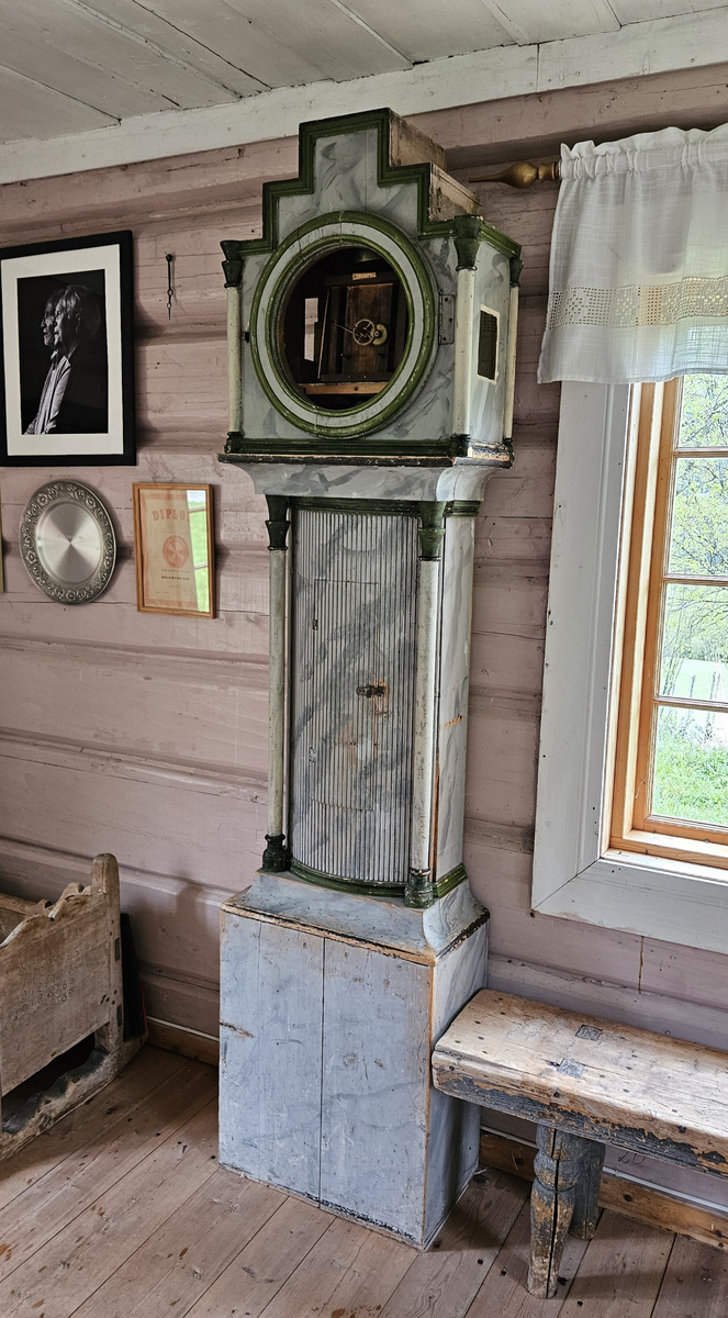 Kasse med dreide hjørnestolper og profilerte lister. Dør til lodd og pendel i midtfeltet. Vulstprofil. Glass på hver side av urverket. Grå marmorering, hvite og grønne og svarte kanter og lister. Foran tallskiva er det glassdør-sirkelformet, glasset mangler. Tallskive av tre, malt - svart romertall. På tallskiva rester av malte menneskefigurer i rødt, grønt, gult, blått. På toppen av kassa fem hull, formodentlig til å feste pyntekuler eller tilsvarende.