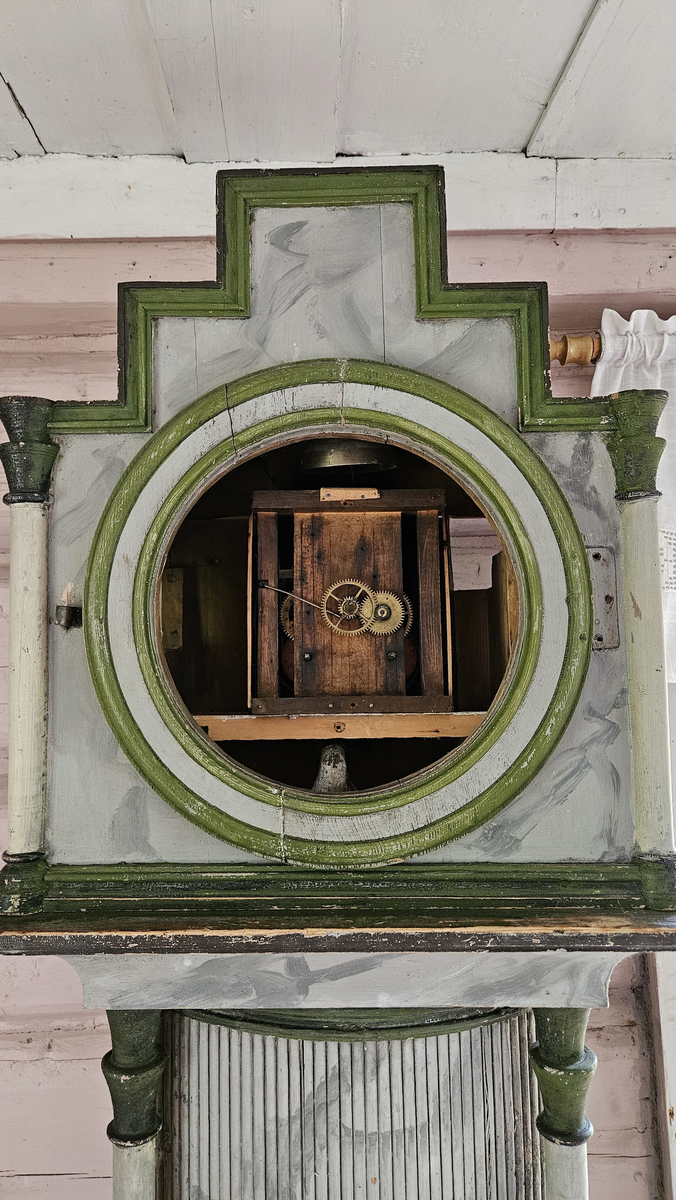 Kasse med dreide hjørnestolper og profilerte lister. Dør til lodd og pendel i midtfeltet. Vulstprofil. Glass på hver side av urverket. Grå marmorering, hvite og grønne og svarte kanter og lister. Foran tallskiva er det glassdør-sirkelformet, glasset mangler. Tallskive av tre, malt - svart romertall. På tallskiva rester av malte menneskefigurer i rødt, grønt, gult, blått. På toppen av kassa fem hull, formodentlig til å feste pyntekuler eller tilsvarende.