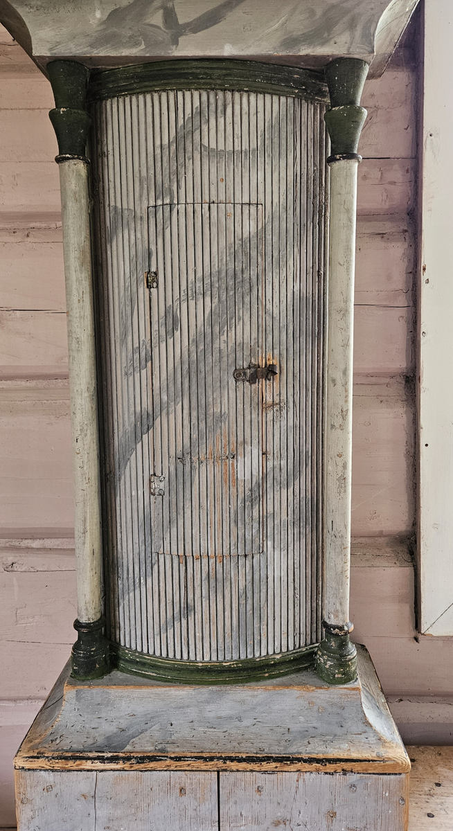 Kasse med dreide hjørnestolper og profilerte lister. Dør til lodd og pendel i midtfeltet. Vulstprofil. Glass på hver side av urverket. Grå marmorering, hvite og grønne og svarte kanter og lister. Foran tallskiva er det glassdør-sirkelformet, glasset mangler. Tallskive av tre, malt - svart romertall. På tallskiva rester av malte menneskefigurer i rødt, grønt, gult, blått. På toppen av kassa fem hull, formodentlig til å feste pyntekuler eller tilsvarende.