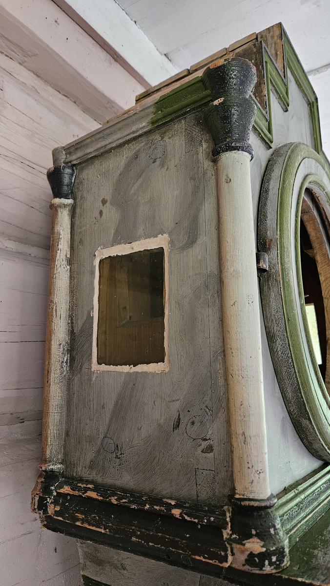 Kasse med dreide hjørnestolper og profilerte lister. Dør til lodd og pendel i midtfeltet. Vulstprofil. Glass på hver side av urverket. Grå marmorering, hvite og grønne og svarte kanter og lister. Foran tallskiva er det glassdør-sirkelformet, glasset mangler. Tallskive av tre, malt - svart romertall. På tallskiva rester av malte menneskefigurer i rødt, grønt, gult, blått. På toppen av kassa fem hull, formodentlig til å feste pyntekuler eller tilsvarende.