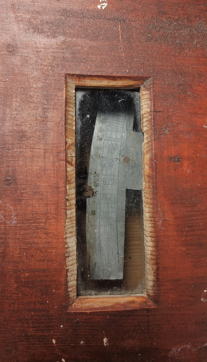 Nivelleringsinstrument bestående av trekasse med glassvindu for avlesing av mål. Fra skiva med mål henger et lodd. På baksida av trekassa er en bøyle; sannsynligvis for feste på et stativ.