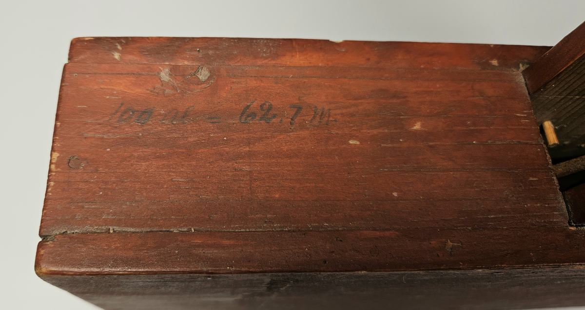 Nivelleringsinstrument bestående av trekasse med glassvindu for avlesing av mål. Fra skiva med mål henger et lodd. På baksida av trekassa er en bøyle; sannsynligvis for feste på et stativ.